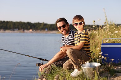 Photo of Happy father and his little son in sunglasses fishing together near river outdoors