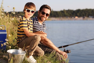 Photo of Happy father and his little son in sunglasses fishing together near river outdoors