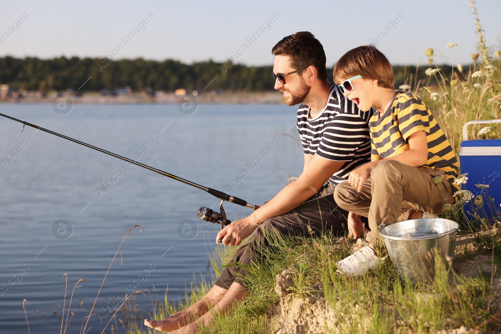 Photo of Happy father and his little son in sunglasses fishing together near river outdoors