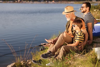 Photo of Smiling grandfather, father and his little son fishing together near river outdoors
