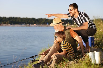 Photo of Smiling grandfather, father and his little son fishing together near river outdoors
