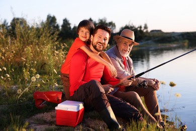 Photo of Smiling grandfather, father and his little son fishing together near river outdoors