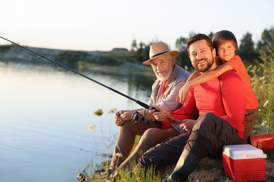 Photo of Smiling grandfather, father and his little son fishing together near river outdoors