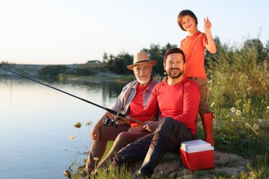Photo of Smiling grandfather, father and his little son fishing together near river outdoors