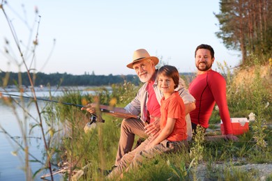 Photo of Smiling grandfather, father and his little son fishing together near river outdoors