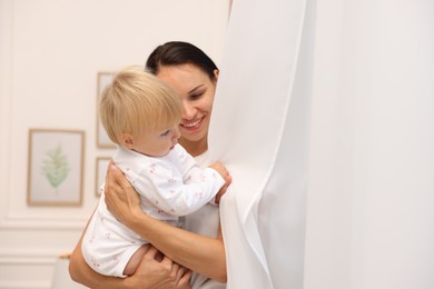 Photo of Smiling mother hugging her cute baby near curtain at home. Space for text