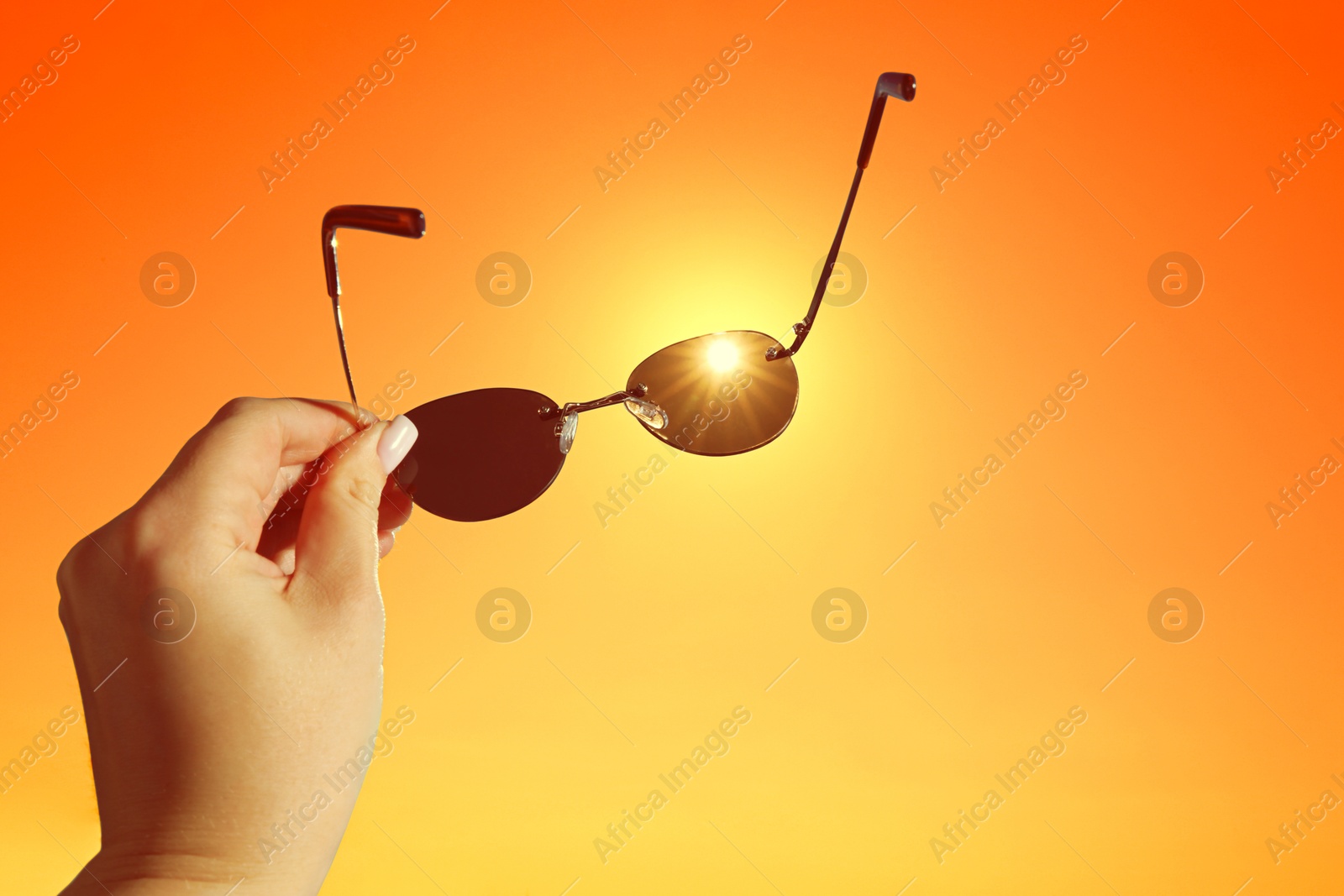 Woman holding sunglasses against orange sky with bright sun, closeup Image of Woman holding sunglasses against orange sky with bright sun, closeup