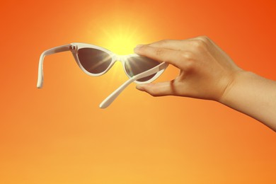 Image of Woman holding sunglasses against orange sky with bright sun, closeup