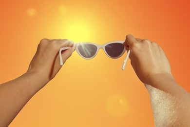 Image of Woman holding sunglasses against orange sky with bright sun, closeup