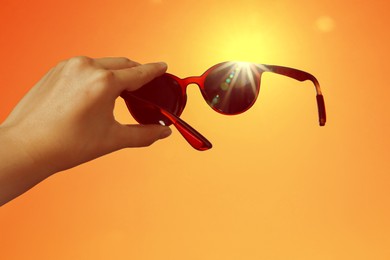 Image of Woman holding sunglasses against orange sky with bright sun, closeup