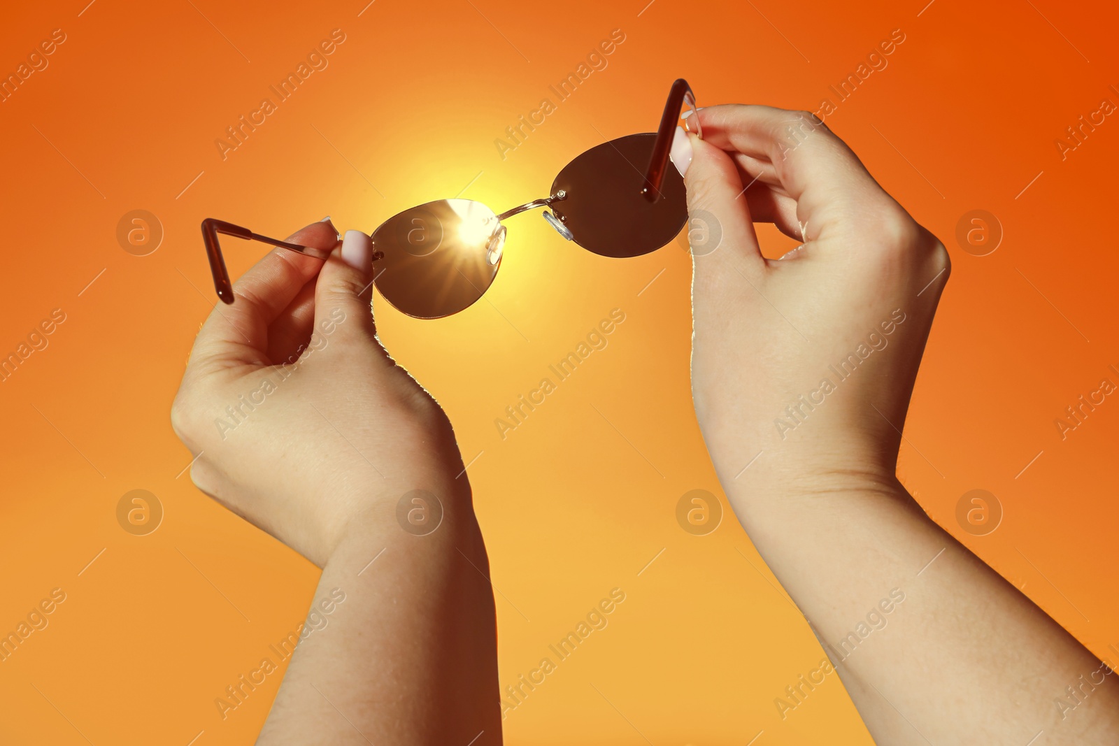 Image of Woman holding sunglasses against orange sky with bright sun, closeup