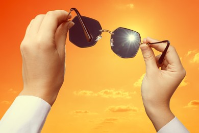 Image of Woman holding sunglasses against orange sky with bright sun, closeup