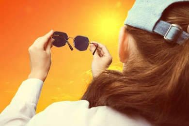 Image of Woman holding sunglasses against orange sky with bright sun, closeup