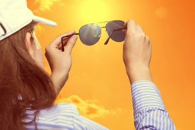 Image of Woman holding sunglasses against orange sky with bright sun, closeup
