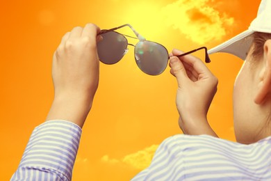 Image of Woman holding sunglasses against orange sky with bright sun, closeup
