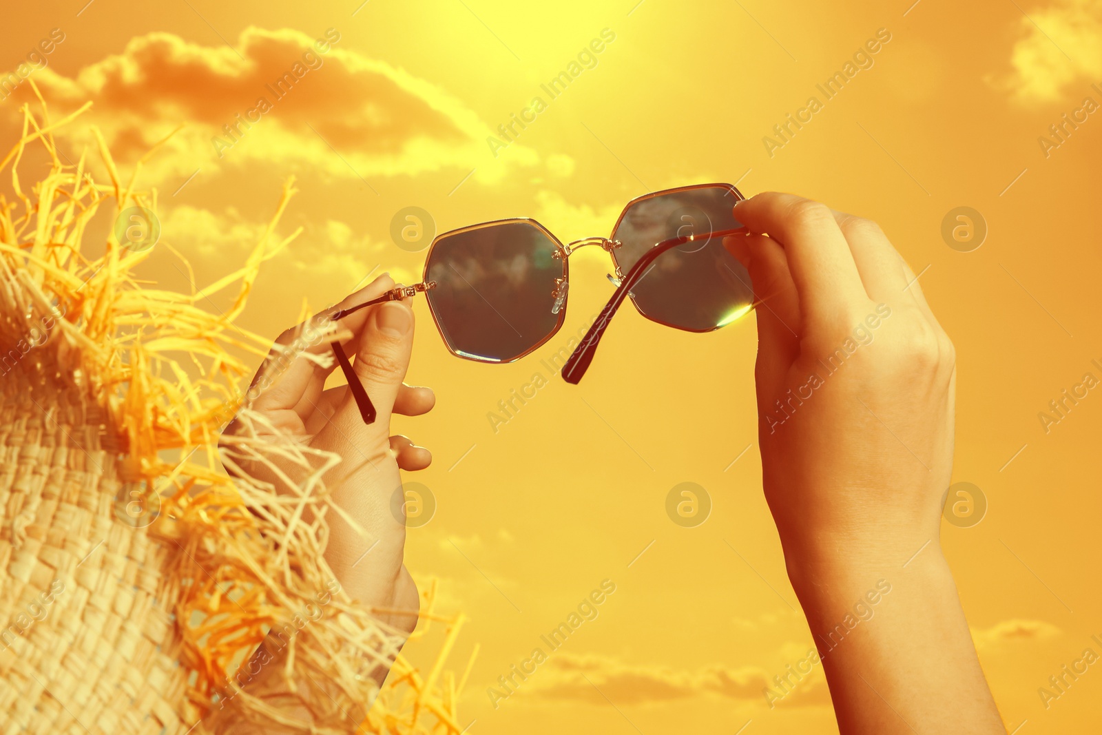 Image of Woman holding sunglasses against orange sky with bright sun, closeup