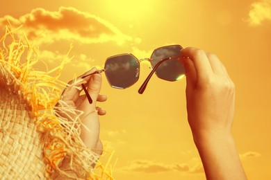 Image of Woman holding sunglasses against orange sky with bright sun, closeup