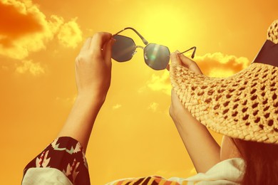 Image of Woman holding sunglasses against orange sky with bright sun, closeup