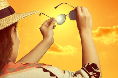 Image of Woman holding sunglasses against orange sky with bright sun, closeup