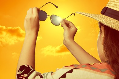 Image of Woman holding sunglasses against orange sky with bright sun, closeup