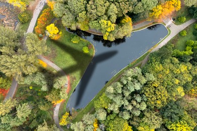 Image of Flying airplane casting shadow on beautiful park with artificial pond, top view