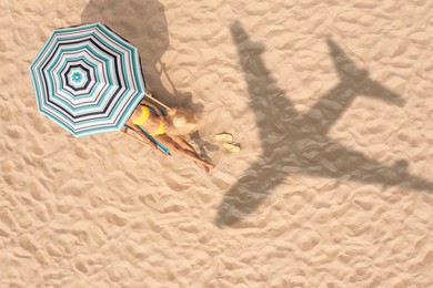 Image of Woman resting under striped umbrella on beach, top view. Flying airplane casting shadow on sand