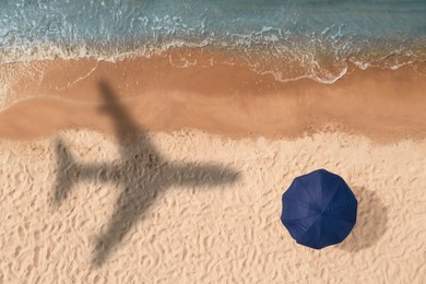 Image of Flying airplane casting shadow on sand with beach umbrella, top view