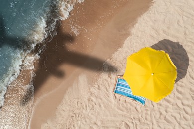 Image of Yellow umbrella and deck chair on beach, top view. Flying airplane casting shadow on sand and water