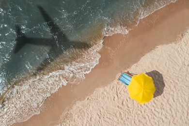 Image of Yellow umbrella and deck chair on beach, top view. Flying airplane casting shadow on sand and water