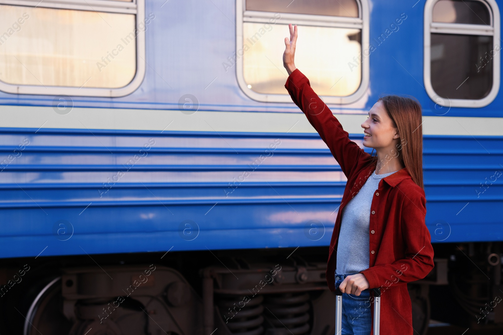 Photo of Goodbye. Happy woman with luggage waving near train outdoors