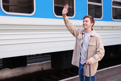 Photo of Goodbye. Happy man with luggage waving near train outdoors, space for text