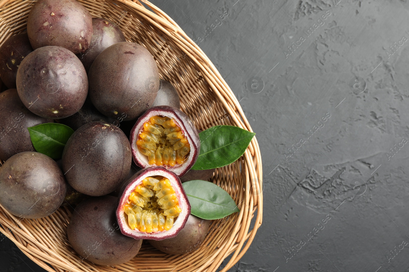 Photo of Fresh passion fruits with green leaves in wicker basket on grey textured table, top view. Space for text