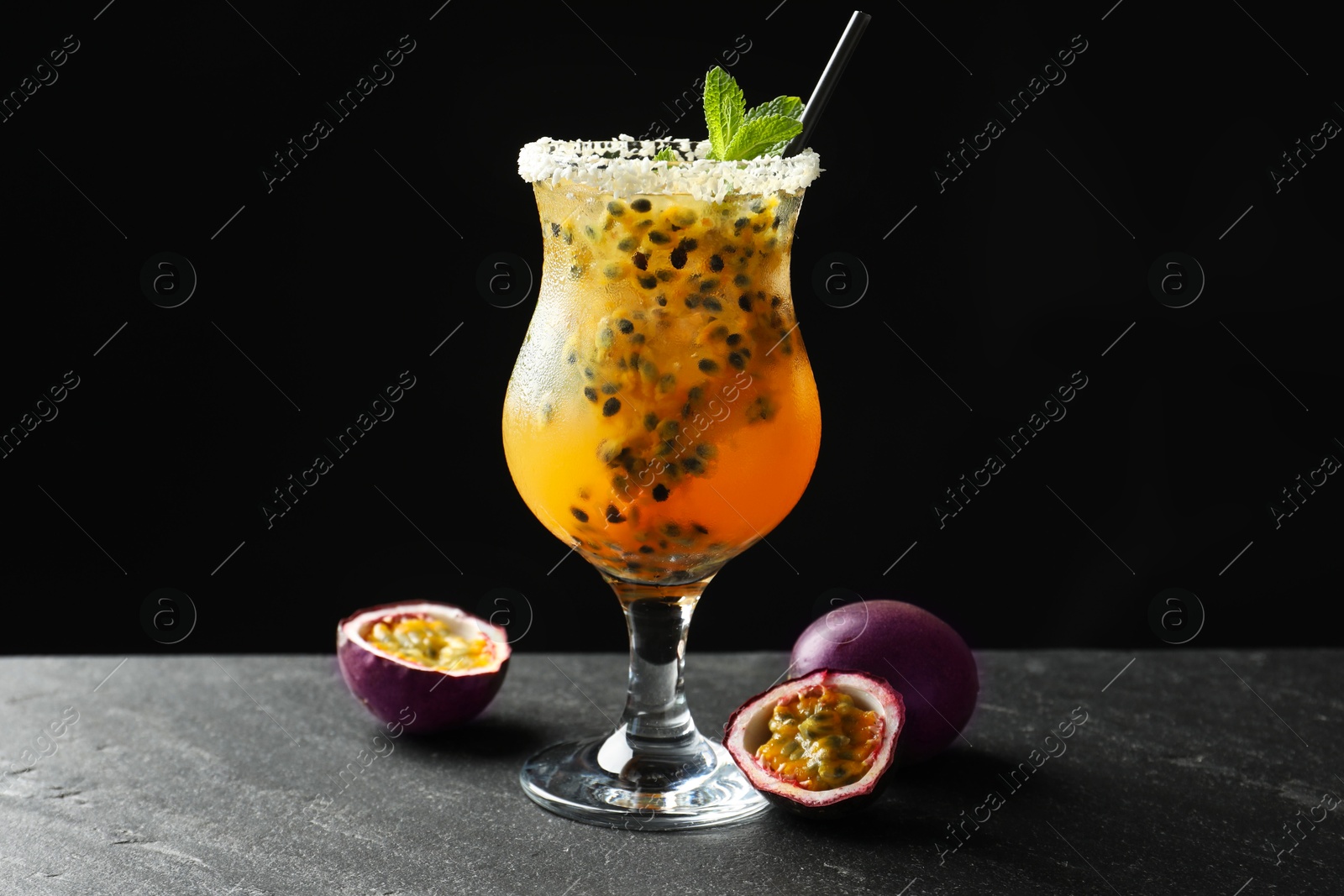 Photo of Tasty passion fruit drink with mint in glass and fresh fruits on dark gray textured table against black background