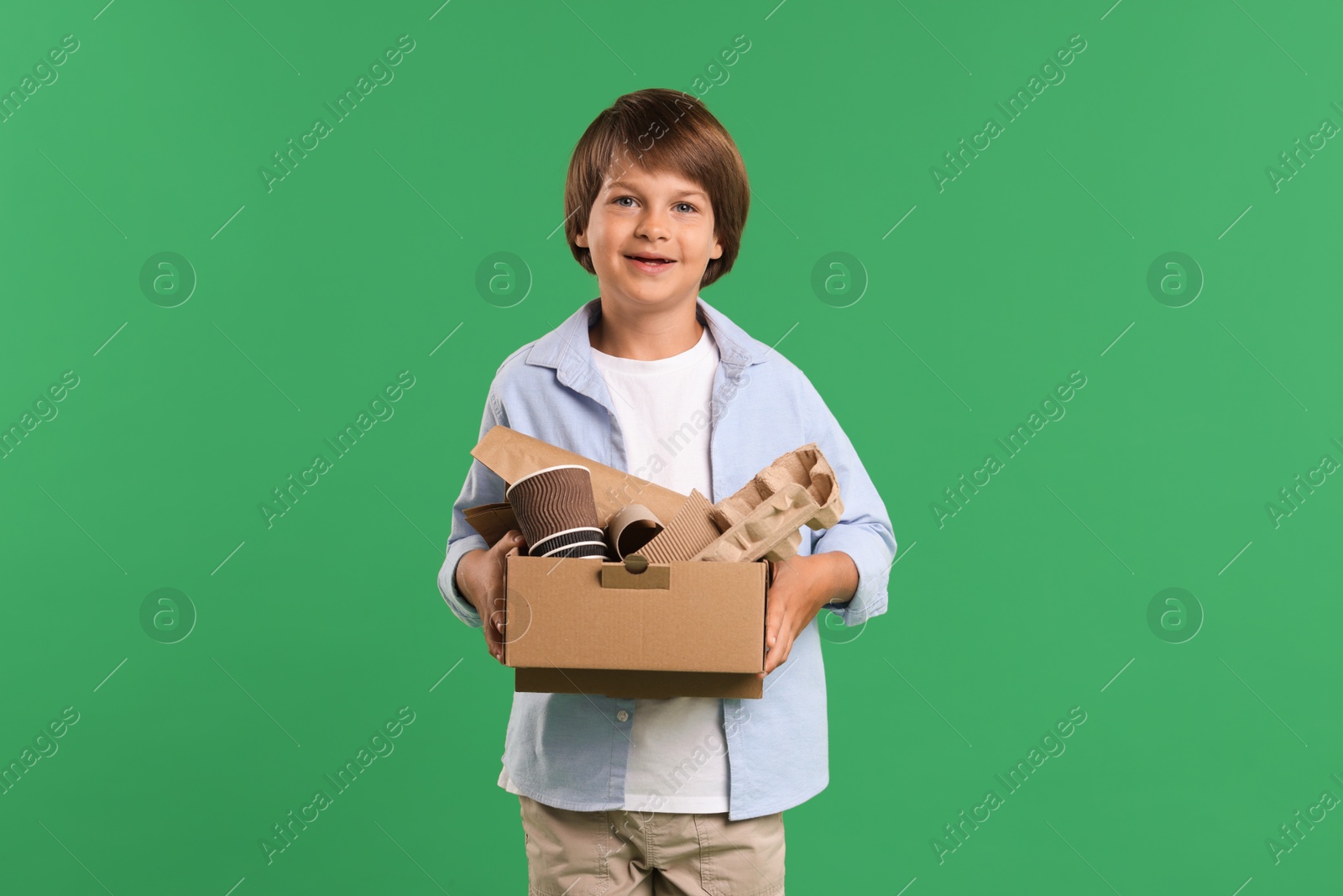 Photo of Recycling. Cute little boy with cardboard box full of different waste paper on green background