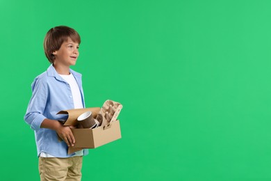 Photo of Recycling. Cute little boy with cardboard box full of different waste paper on green background, space for text