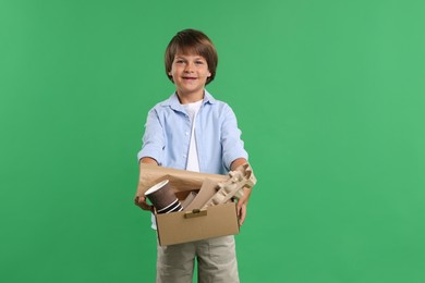 Photo of Recycling. Cute little boy with cardboard box full of different waste paper on green background