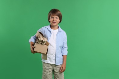 Photo of Recycling. Cute little boy with cardboard box full of different waste paper on green background