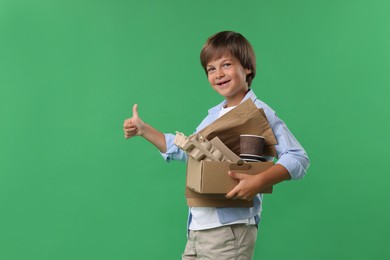 Photo of Recycling. Cute little boy with cardboard box full of different waste paper showing thumbs up on green background
