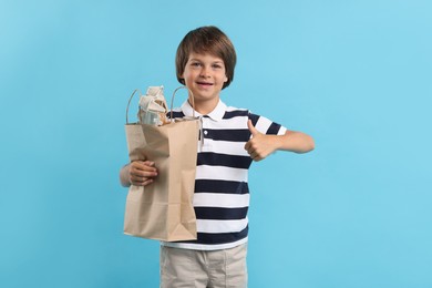 Photo of Recycling. Cute little boy with bag full of different waste paper showing thumbs up on light blue background