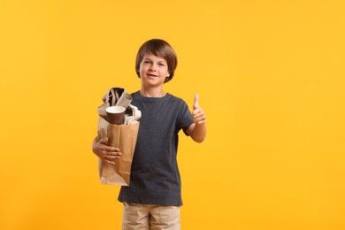 Photo of Recycling. Cute little boy with bag full of different waste paper showing thumbs up on orange background