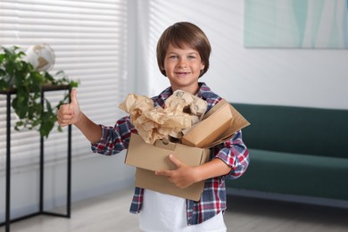Photo of Recycling. Cute little boy with cardboard box full of different waste paper showing thumbs up at home
