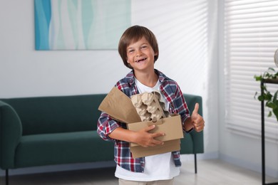Photo of Recycling. Cute little boy with cardboard box full of different waste paper showing thumbs up at home