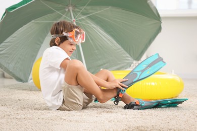 Photo of Dreaming about vacation at sea. Little boy in diving mask wearing flippers near inflatable ring indoors