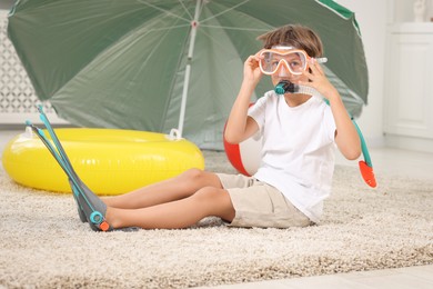 Photo of Dreaming about vacation at sea. Little boy in diving mask, snorkel and flippers near inflatable ring indoors