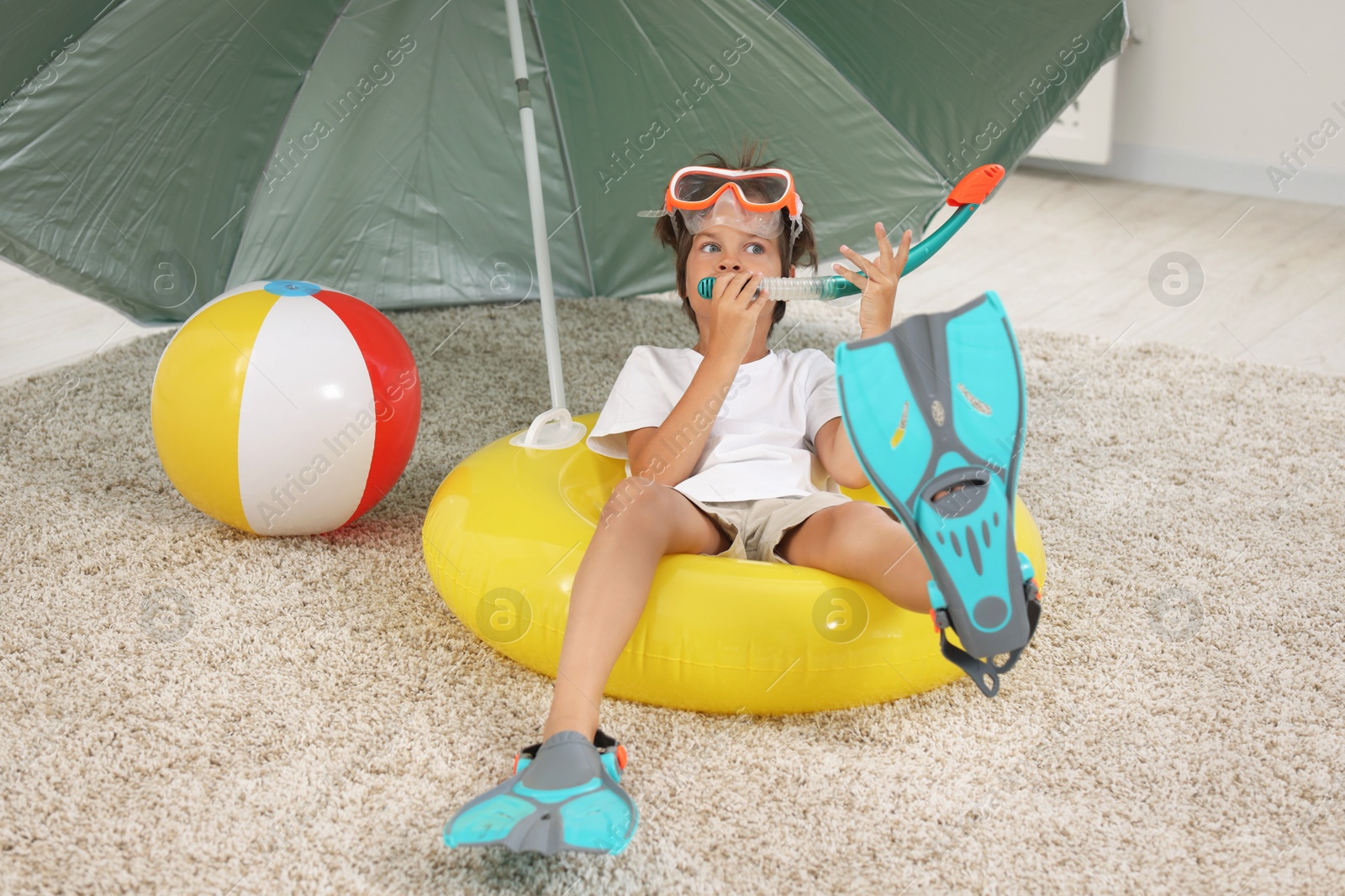 Photo of Dreaming about vacation at sea. Little boy with diving mask, snorkel, flippers and inflatable ring on floor indoors