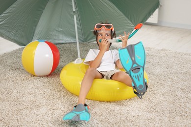 Photo of Dreaming about vacation at sea. Little boy with diving mask, snorkel, flippers and inflatable ring on floor indoors