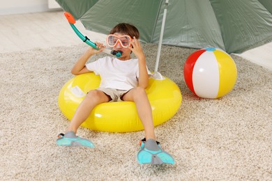 Photo of Dreaming about vacation at sea. Little boy with diving mask, snorkel, flippers and inflatable ring on floor indoors