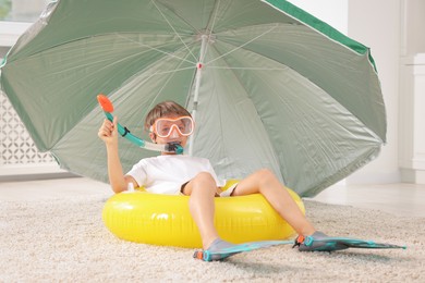 Photo of Dreaming about vacation at sea. Little boy with diving mask, snorkel, flippers and inflatable ring on floor indoors