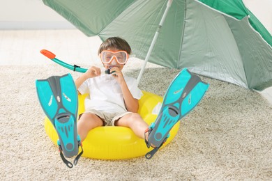 Photo of Dreaming about vacation at sea. Little boy with diving mask, snorkel, flippers and inflatable ring on floor indoors