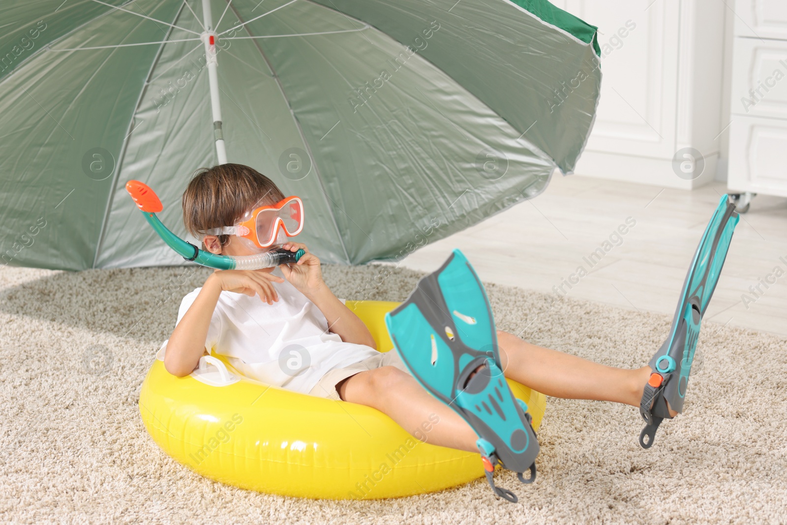 Photo of Dreaming about vacation at sea. Little boy with diving mask, snorkel, flippers and inflatable ring on floor indoors